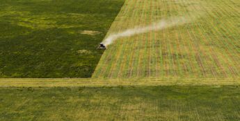 Aerial view of a tractor harvesting a large green field, leaving visible lines and a trail of dust behind.