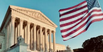 The United States Supreme Court building with an American flag waving prominently in the foreground against a clear sky.