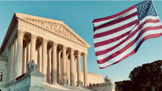 The United States Supreme Court building with an American flag waving prominently in the foreground against a clear sky.