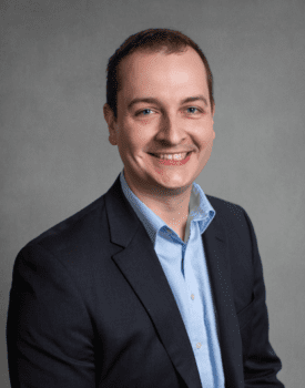 Lane Buchanan, wearing a dark suit jacket and light blue shirt, is smiling at the camera against a plain gray background.