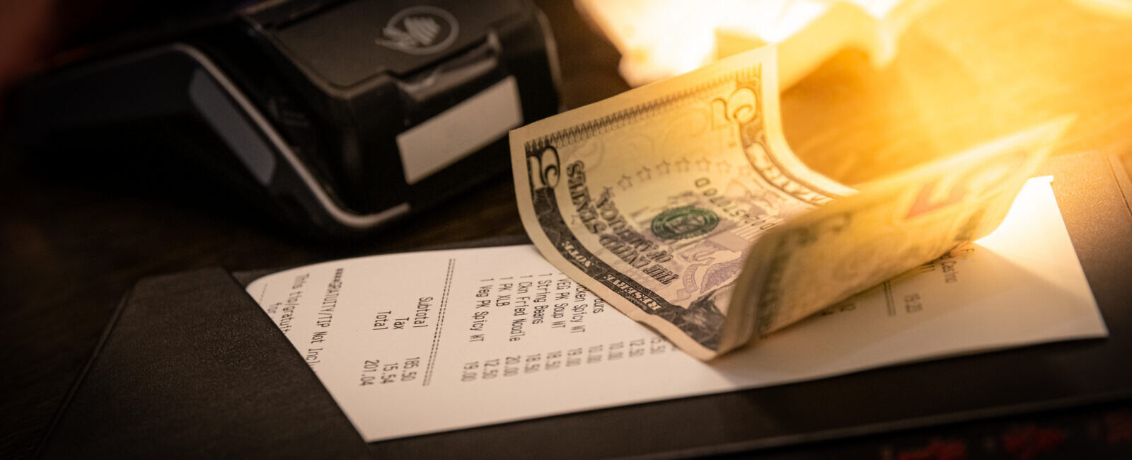 A restaurant bill with a twenty-dollar bill placed on top, next to a payment terminal on a dimly lit table.