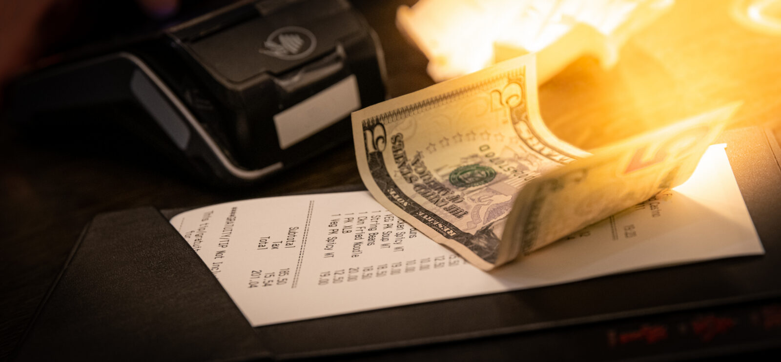 A restaurant bill with a twenty-dollar bill placed on top, next to a payment terminal on a dimly lit table.