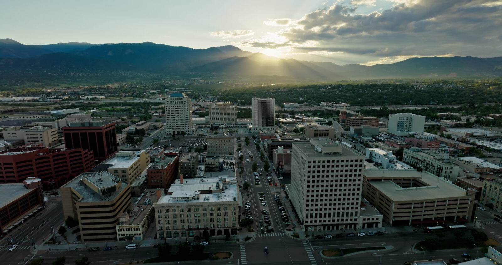 Aerial view of Colorado Springs downtown with multiple office buildings, cars on the streets, and mountains in the background at sunset.