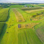Aerial view of a rural landscape with green fields, scattered hay bales, farm buildings, and a forested area on the left side—reflecting the precision found in ccreditation by the ANSI National Accreditation Board (ANAB) to ISO/IEC 17021‑1.