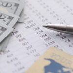A silver pen, several US $100 bills, and a bank card rest on financial documents with numbers and data tables.