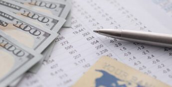 A silver pen, several US $100 bills, and a bank card rest on financial documents with numbers and data tables.