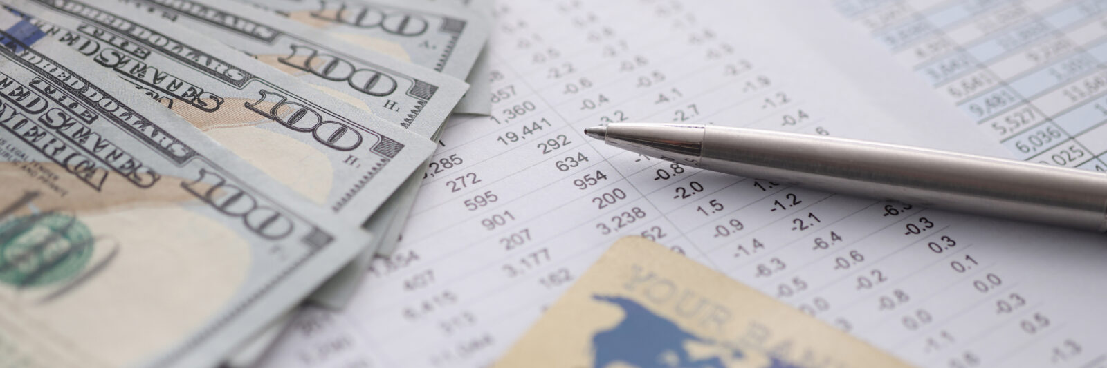 A silver pen, several US $100 bills, and a bank card rest on financial documents with numbers and data tables.