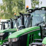 A row of new green tractors with large tires and orange lights is parked outdoors on a sunny day.