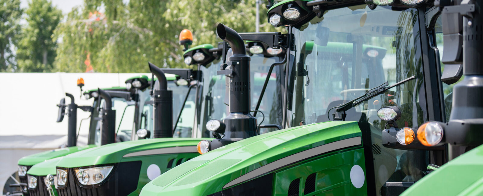 A row of new green tractors with large tires and orange lights is parked outdoors on a sunny day.