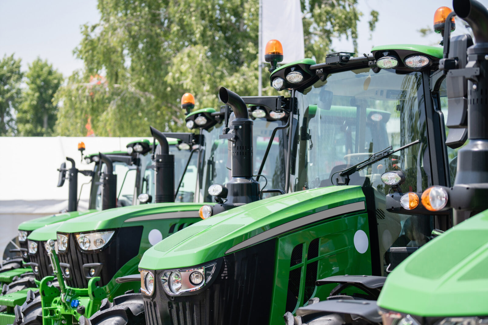 A row of new green tractors with large tires and orange lights is parked outdoors on a sunny day.