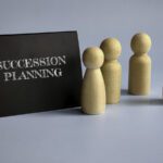 A black sign with "Succession Planning" in white letters stands beside four wooden human-shaped figures, symbolizing Leadership Transitions, against a light blue background.