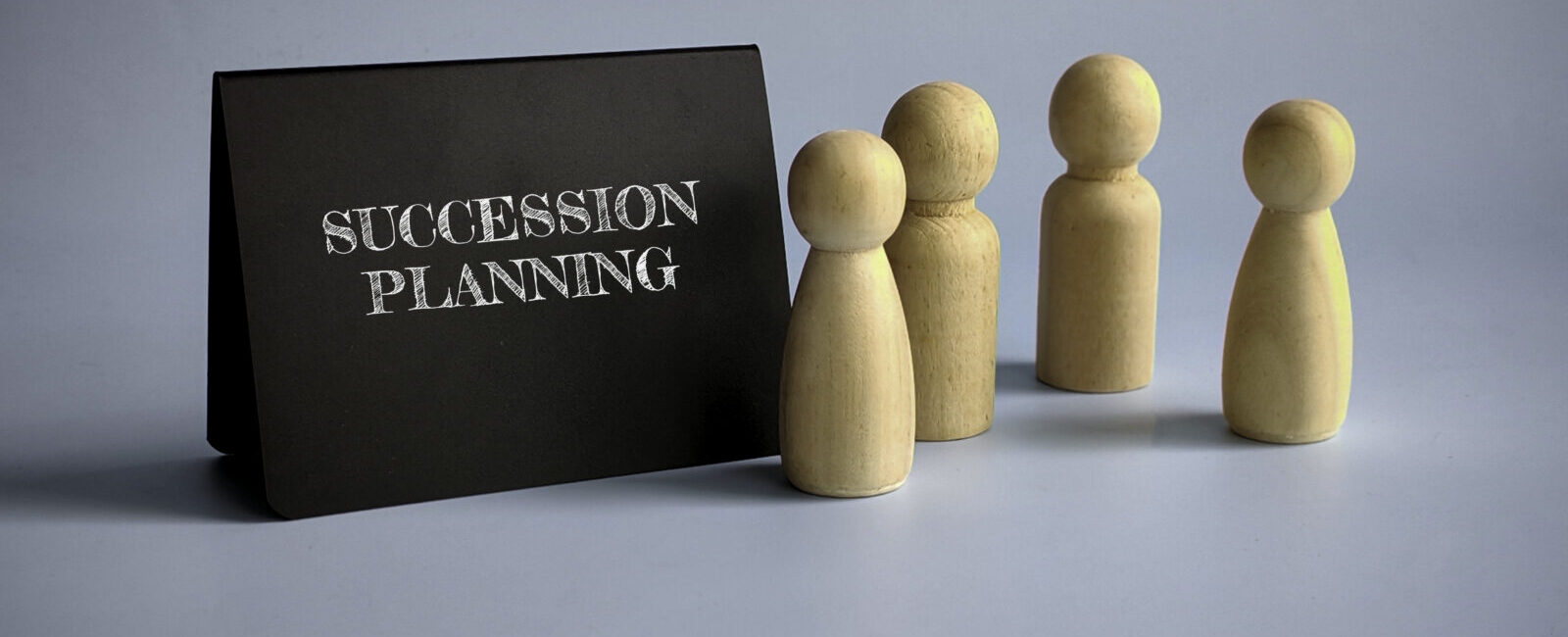 A black sign with "Succession Planning" in white letters stands beside four wooden human-shaped figures, symbolizing Leadership Transitions, against a light blue background.