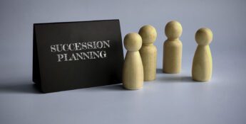 A black sign with "Succession Planning" in white letters stands beside four wooden human-shaped figures, symbolizing Leadership Transitions, against a light blue background.