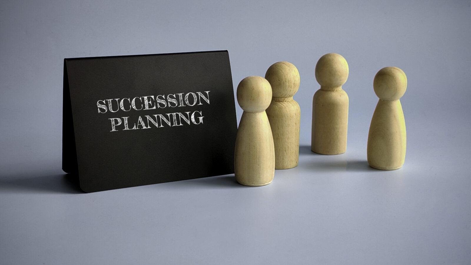 A black sign with "Succession Planning" in white letters stands beside four wooden human-shaped figures, symbolizing Leadership Transitions, against a light blue background.