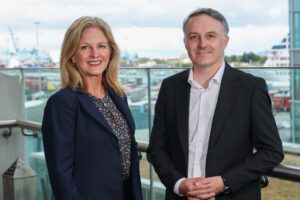 Two people in business attire stand on a balcony with a marina and shipping containers visible in the background.