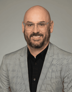 A man with a bald head, beard, and clear glasses wears a gray plaid blazer over a black shirt, posing in front of a plain gray background.