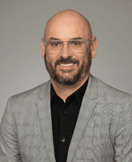 A man with a bald head, beard, and clear glasses wears a gray plaid blazer over a black shirt, posing in front of a plain gray background.