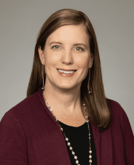 A woman with straight, shoulder-length light brown hair wearing a maroon cardigan, black top, and beaded necklace, smiling at the camera against a plain background.