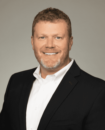 A man with short, light brown hair and a beard wears a white dress shirt and black blazer, smiling in front of a plain light gray background.