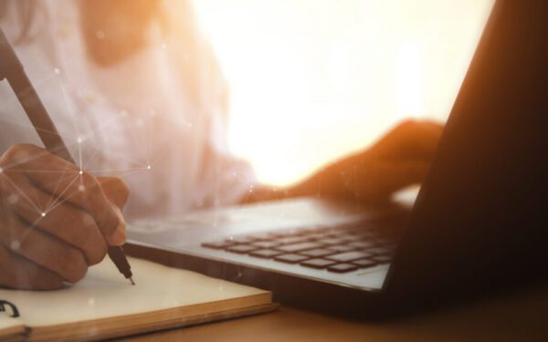 Person writes in a notebook with a pen while using a laptop at a desk. Sunlight shines in the background.
