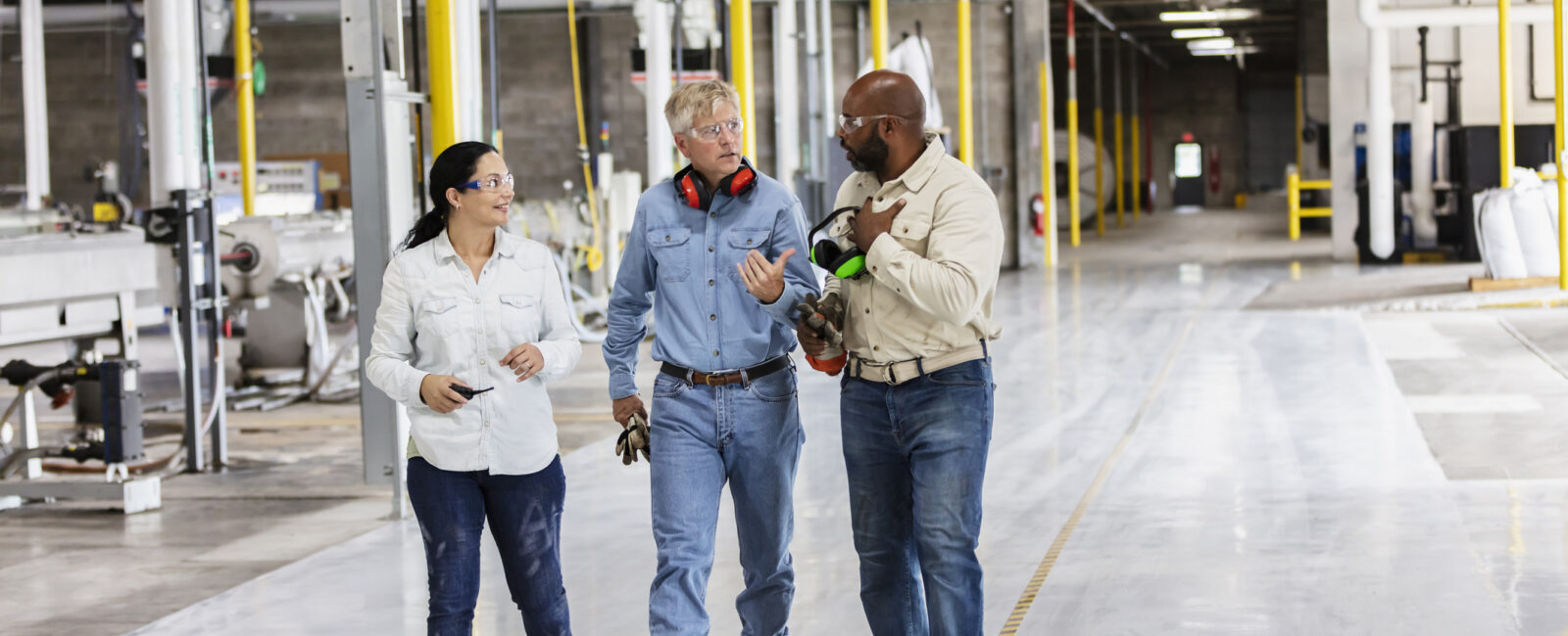 Three people in work attire walk and talk in a large, well-lit industrial facility with yellow pillars and equipment visible in the background.
