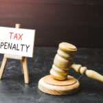 A wooden gavel rests beside a small easel displaying a sign that reads "TAX PENALTY" on a dark surface with a black background, subtly highlighting the significance of understanding options like the IRS Automatic Exemption From Penalty (AEP) for those seeking relief.