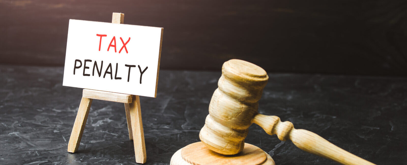 A wooden gavel rests beside a small easel displaying a sign that reads "TAX PENALTY" on a dark surface with a black background, subtly highlighting the significance of understanding options like the IRS Automatic Exemption From Penalty (AEP) for those seeking relief.