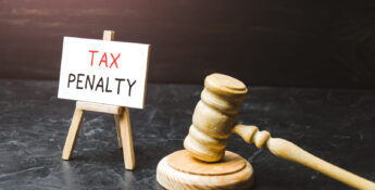 A wooden gavel rests beside a small easel displaying a sign that reads "TAX PENALTY" on a dark surface with a black background, subtly highlighting the significance of understanding options like the IRS Automatic Exemption From Penalty (AEP) for those seeking relief.