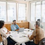 Four people sit around a table in a modern office, engaged in a meeting with laptops, notebooks, and paperwork visible—a setting where peer groups are leadership think tanks, fostering collaboration and innovative problem-solving.