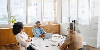 Four people sit around a table in a modern office, engaged in a meeting with laptops, notebooks, and paperwork visible—a setting where peer groups are leadership think tanks, fostering collaboration and innovative problem-solving.