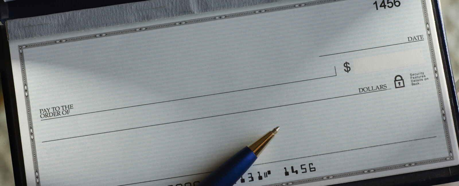 A blank check in a checkbook with a pen resting on it, ready to be written, showing check number 1456 and account details at the bottom—a subtle reminder of how easily such financial tools can contribute to check fraud losses if not handled securely.