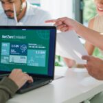 Four people sit at a table discussing papers; a laptop screen displays a Net Zero Emission infographic featuring charts and statistics about clean energy, sustainability, and the latest updates from the SBTi Net-Zero Standard V2.0.