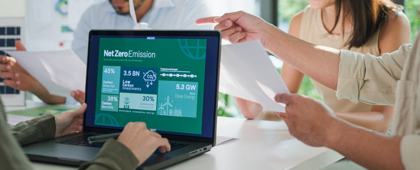 Four people sit at a table discussing papers; a laptop screen displays a Net Zero Emission infographic featuring charts and statistics about clean energy, sustainability, and the latest updates from the SBTi Net-Zero Standard V2.0.