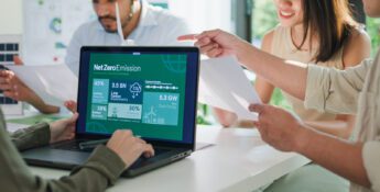 Four people sit at a table discussing papers; a laptop screen displays a Net Zero Emission infographic featuring charts and statistics about clean energy, sustainability, and the latest updates from the SBTi Net-Zero Standard V2.0.
