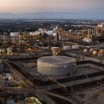Aerial view of an industrial oil refinery complex with storage tanks, pipelines, and city in the background at sunset.