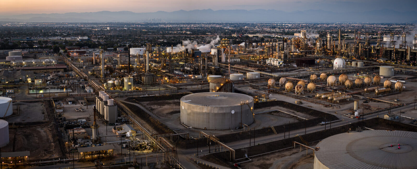 Aerial view of an industrial oil refinery complex with storage tanks, pipelines, and city in the background at sunset.