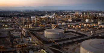 Aerial view of an industrial oil refinery complex with storage tanks, pipelines, and city in the background at sunset.