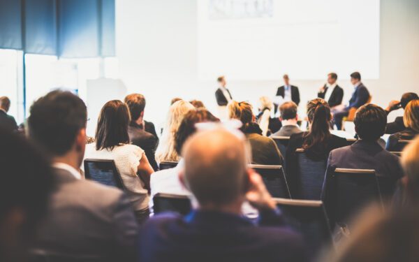 Audience seated in a conference room, watching the TCA Client Corner panel with four speakers on stage and a presentation projected behind them.