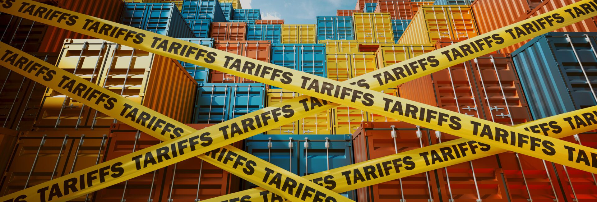 Stacks of colorful shipping containers are blocked by yellow tape labeled “TARIFFS,” symbolizing trade barriers. Blue sky and scattered clouds are visible above the containers.