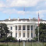 The White House in Washington, D.C., viewed from the front lawn with trees, a flagpole, and people visible in the background.