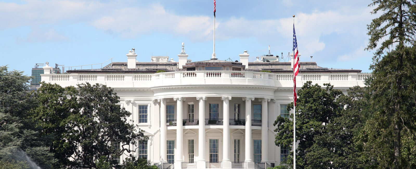 The White House in Washington, D.C., viewed from the front lawn with trees, a flagpole, and people visible in the background.