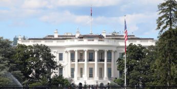 The White House in Washington, D.C., viewed from the front lawn with trees, a flagpole, and people visible in the background.
