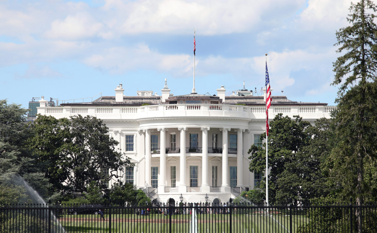 The White House in Washington, D.C., viewed from the front lawn with trees, a flagpole, and people visible in the background.