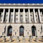 Large neoclassical government building with tall columns, arched doorways, numerous windows, and several small potted plants arranged in a row in front—an imposing structure that could easily serve as the backdrop for important announcements, such as when the IRS increases standard mileage rates.