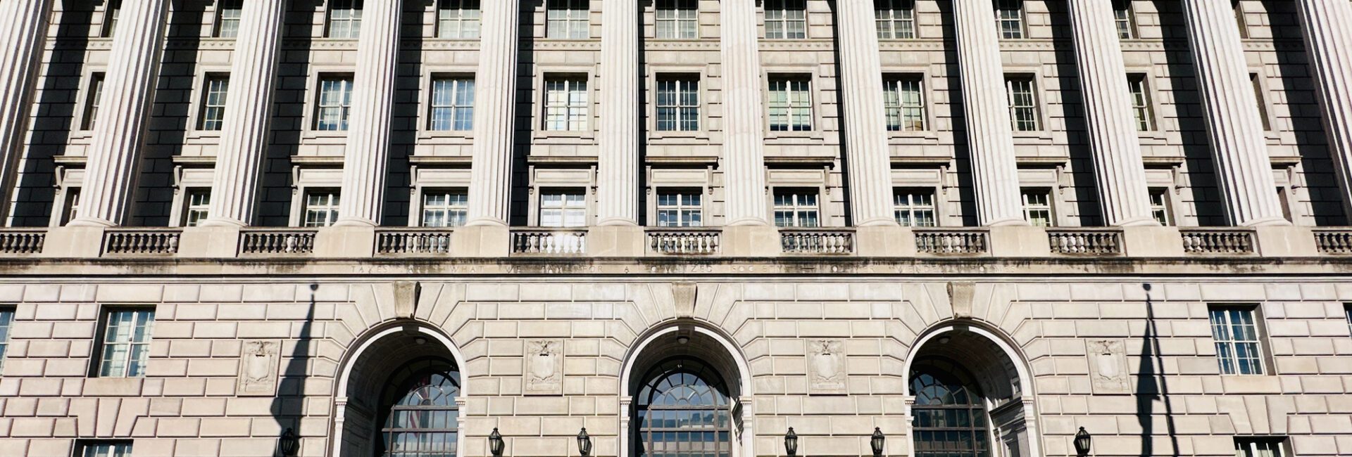 Large neoclassical government building with tall columns, arched doorways, numerous windows, and several small potted plants arranged in a row in front—an imposing structure that could easily serve as the backdrop for important announcements, such as when the IRS increases standard mileage rates.