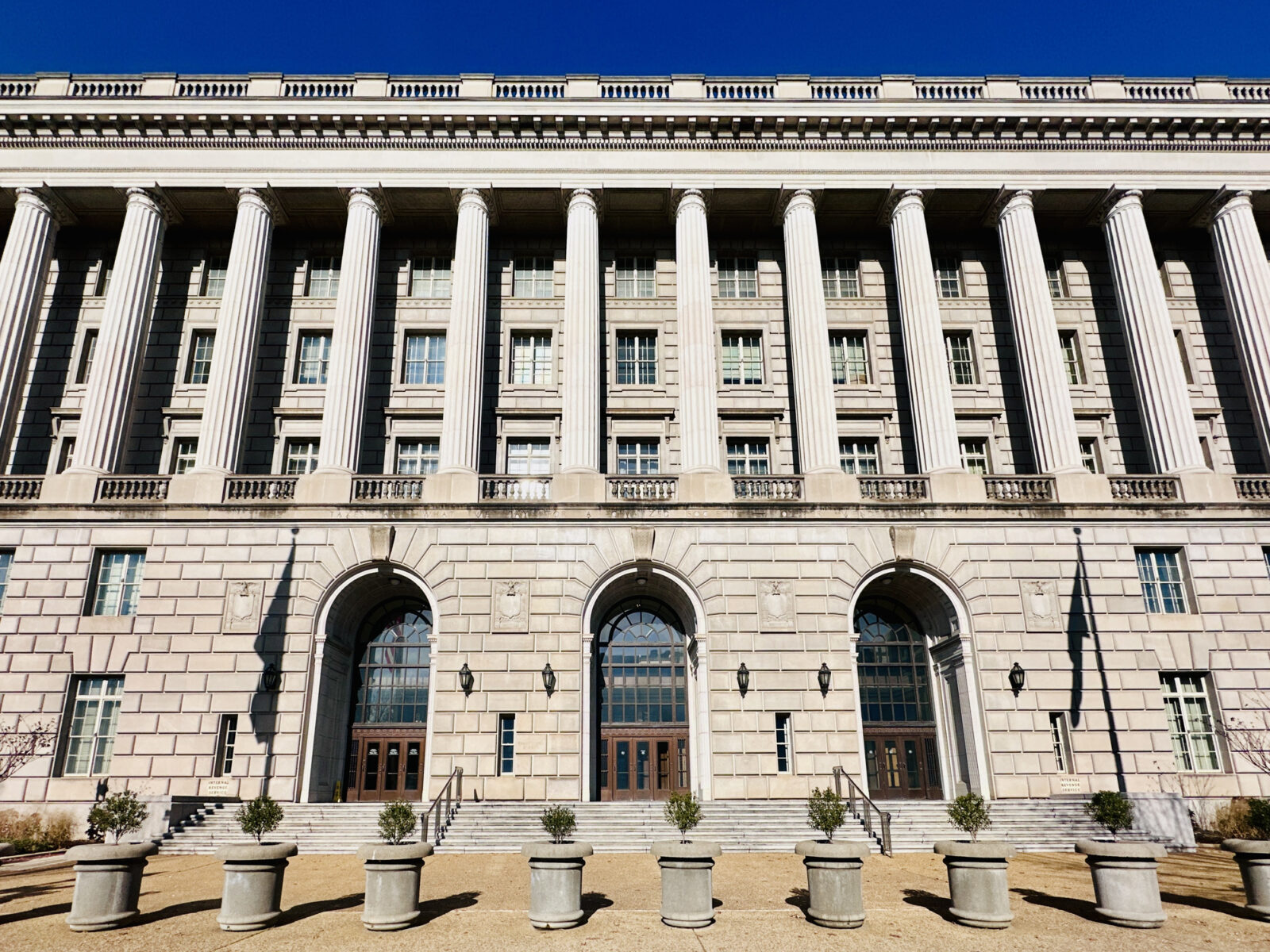 Large neoclassical government building with tall columns, arched doorways, numerous windows, and several small potted plants arranged in a row in front—an imposing structure that could easily serve as the backdrop for important announcements, such as when the IRS increases standard mileage rates.
