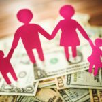 Pink paper cutouts of a family of four holding hands stand on a pile of assorted U.S. dollar bills spread out on a wooden surface, symbolizing the importance of transparency with compensation in supporting families’ financial well-being.