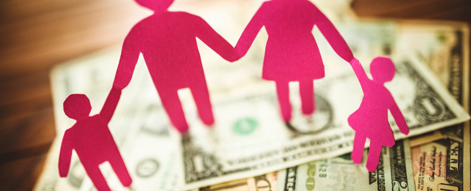 Pink paper cutouts of a family of four holding hands stand on a pile of assorted U.S. dollar bills spread out on a wooden surface, symbolizing the importance of transparency with compensation in supporting families’ financial well-being.