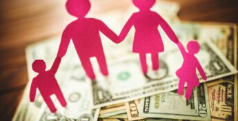 Pink paper cutouts of a family of four holding hands stand on a pile of assorted U.S. dollar bills spread out on a wooden surface, symbolizing the importance of transparency with compensation in supporting families’ financial well-being.