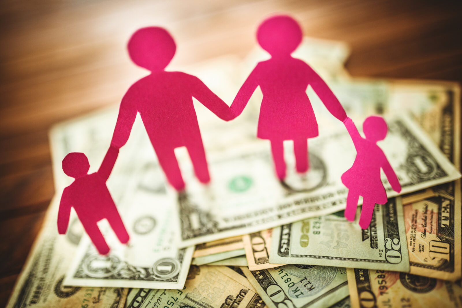 Pink paper cutouts of a family of four holding hands stand on a pile of assorted U.S. dollar bills spread out on a wooden surface, symbolizing the importance of transparency with compensation in supporting families’ financial well-being.